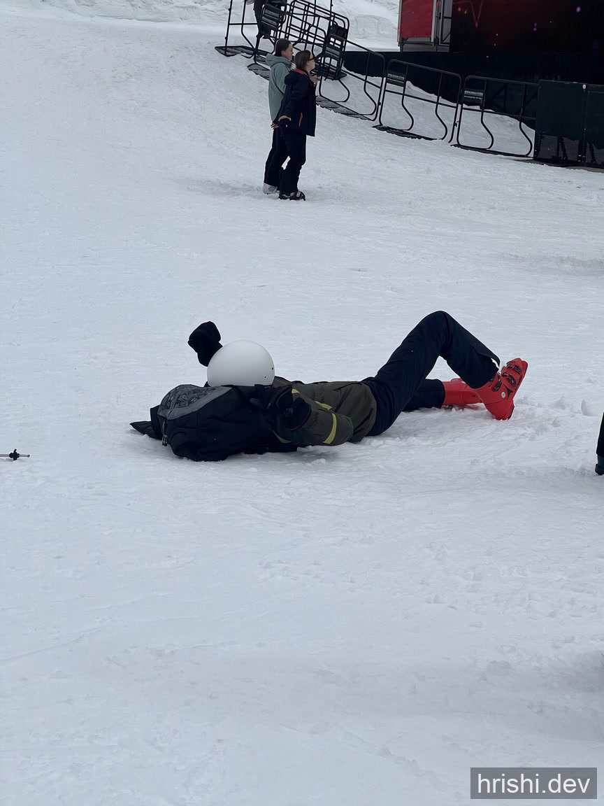 Lying flat on a snowy slope after a fall, with two people looking on in the background